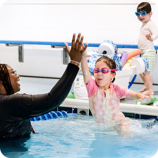 An indoor swimming lesson scene: a child in a pink swim shirt and goggles splashes in the pool and high‑fives an instructor in the water, while another child in blue goggles and swim trunks stands on the deck nearby. Pool equipment like noodles, kickboards, and pull buoys sits on a cart beside the pool, and lane markers float in the water.